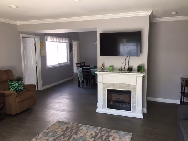 A cozy living room area with a brown armchair on the left, a white electric fireplace with a mounted flat-screen TV above it in the center, and a dining area with a table and chairs visible through an open doorway. The room has gray walls, wood flooring, and a patterned rug in the foreground.