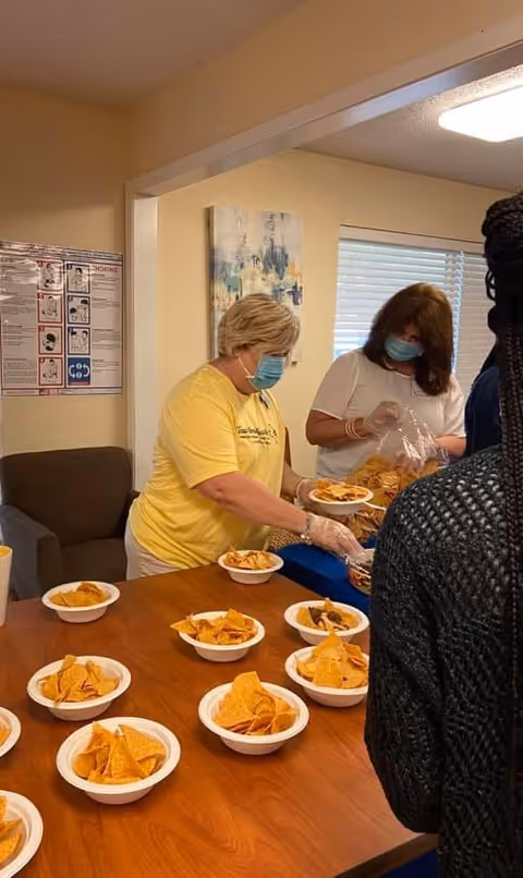 Two masked staff members portion bowls of tortilla chips on a table inside a communal dining area.