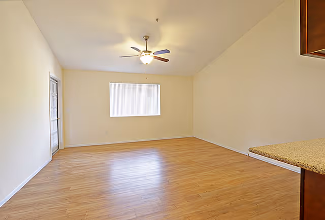 Empty room with light wood flooring, beige walls, a ceiling fan with light, a window with vertical blinds, and a door with glass panels. A granite countertop is partially visible on the right side.