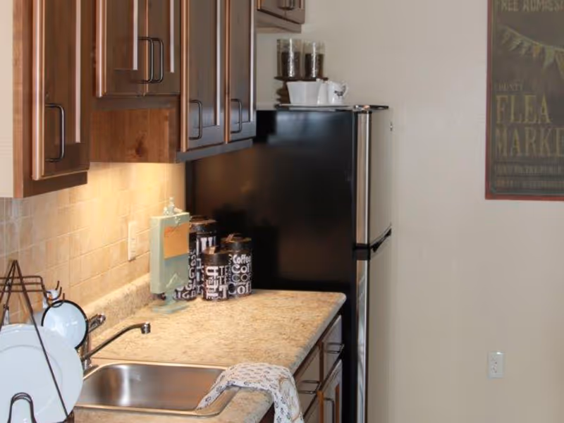 Small kitchen countertop with a sink, dish rack, upper cabinets, coffee canisters, and a stainless refrigerator.