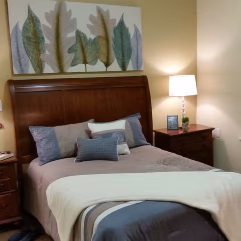 A cozy bedroom with a wooden headboard bed made with multiple pillows and a beige and gray comforter. There is a nightstand on each side of the bed, one with a lamp, a small plant, and a framed photo. Above the bed hangs a large artwork featuring various leaf shapes in muted colors.