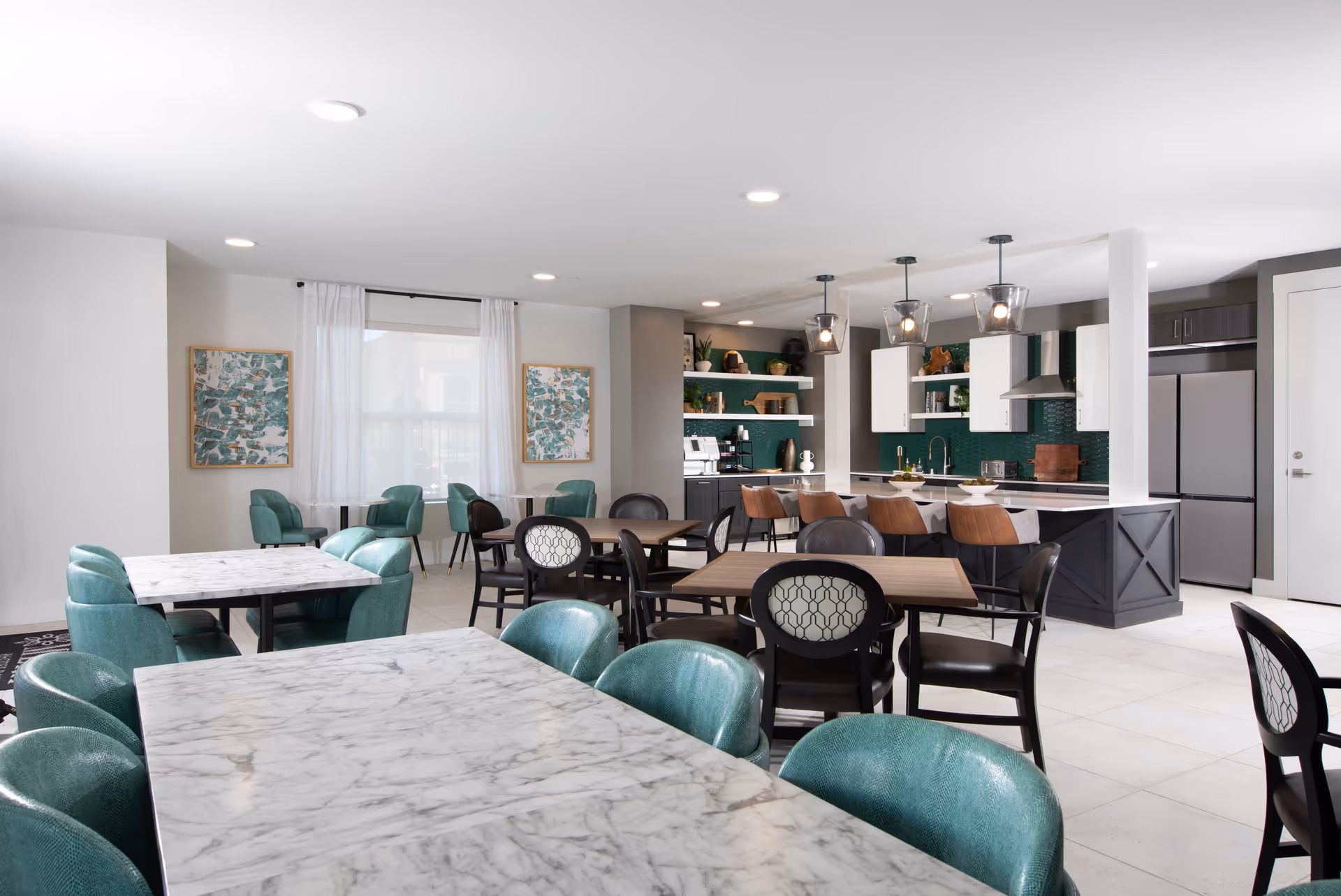 A modern communal dining and kitchen area with marble and wood tables surrounded by teal and black chairs. The kitchen features white cabinets, a green tile backsplash, stainless steel appliances, and three pendant lights hanging over a breakfast bar with brown stools. Two abstract paintings hang on the wall near a window with sheer white curtains.