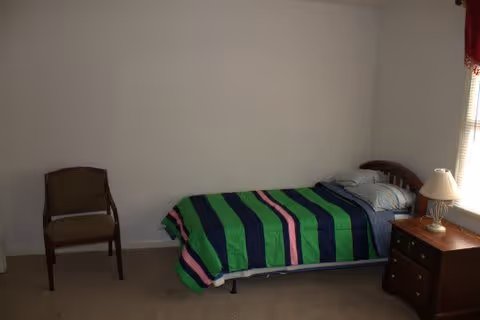 A simple bedroom with a single bed covered with a green, blue, and pink striped blanket. Next to the bed is a wooden nightstand with a lamp on it. There is a wooden chair against the wall and a window with blinds partially open letting in natural light.