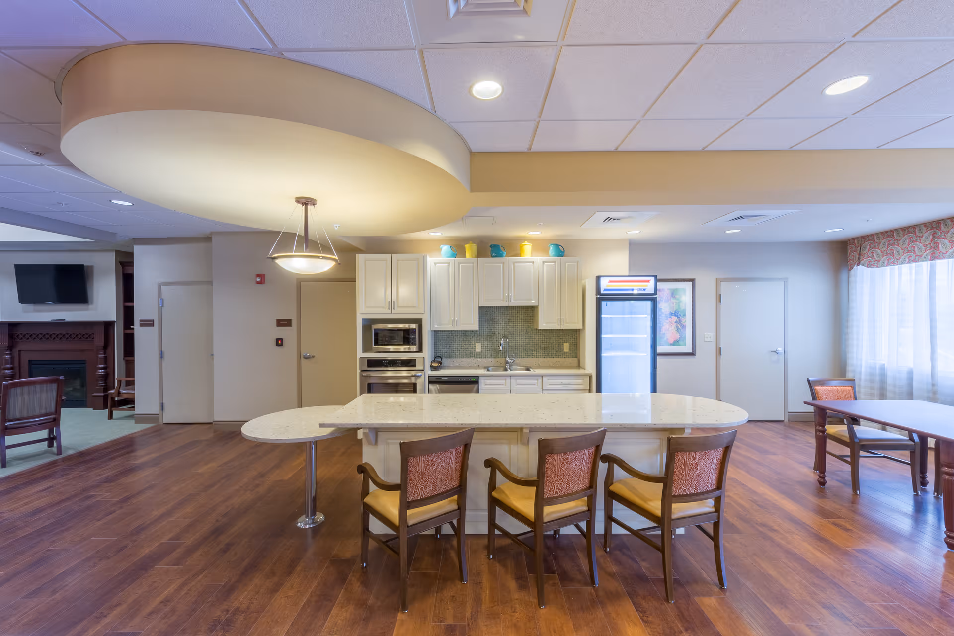 Communal kitchen and dining area with a large island, four chairs, white cabinets, appliances, and hardwood floors.