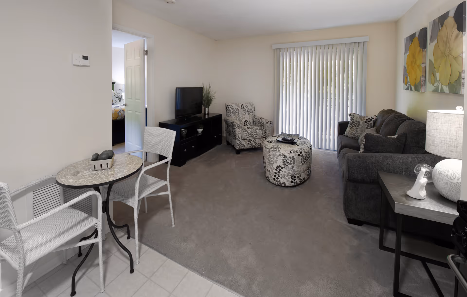 A cozy living room with a gray sofa, patterned armchair, and matching ottoman. A TV sits on a black stand against the wall. There is a small round table with two white chairs on a tiled floor near the entrance. Vertical blinds cover a sliding glass door, and two floral paintings hang on the wall above the sofa. A side table with a lamp and decorative items is next to the sofa.