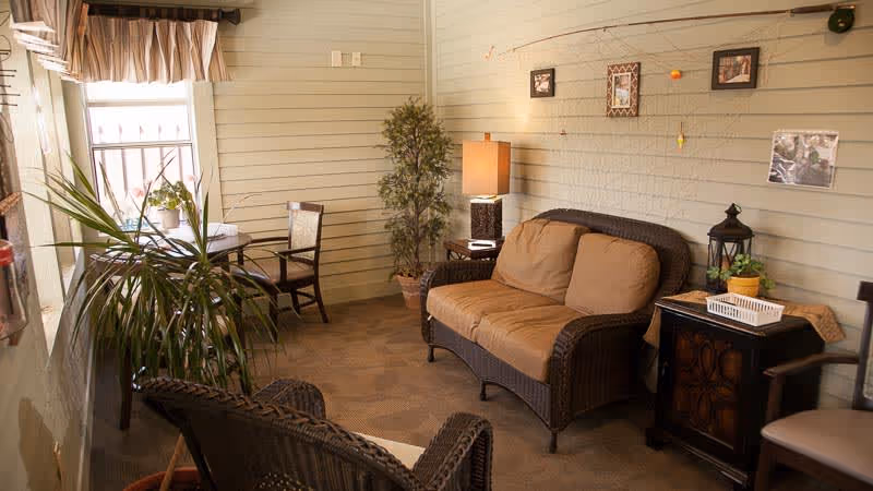 A cozy indoor sitting area with wicker furniture including a cushioned loveseat and chair, a small table with two chairs near a window with a valance, a potted plant, a lamp on a side table, and framed pictures on the wall.