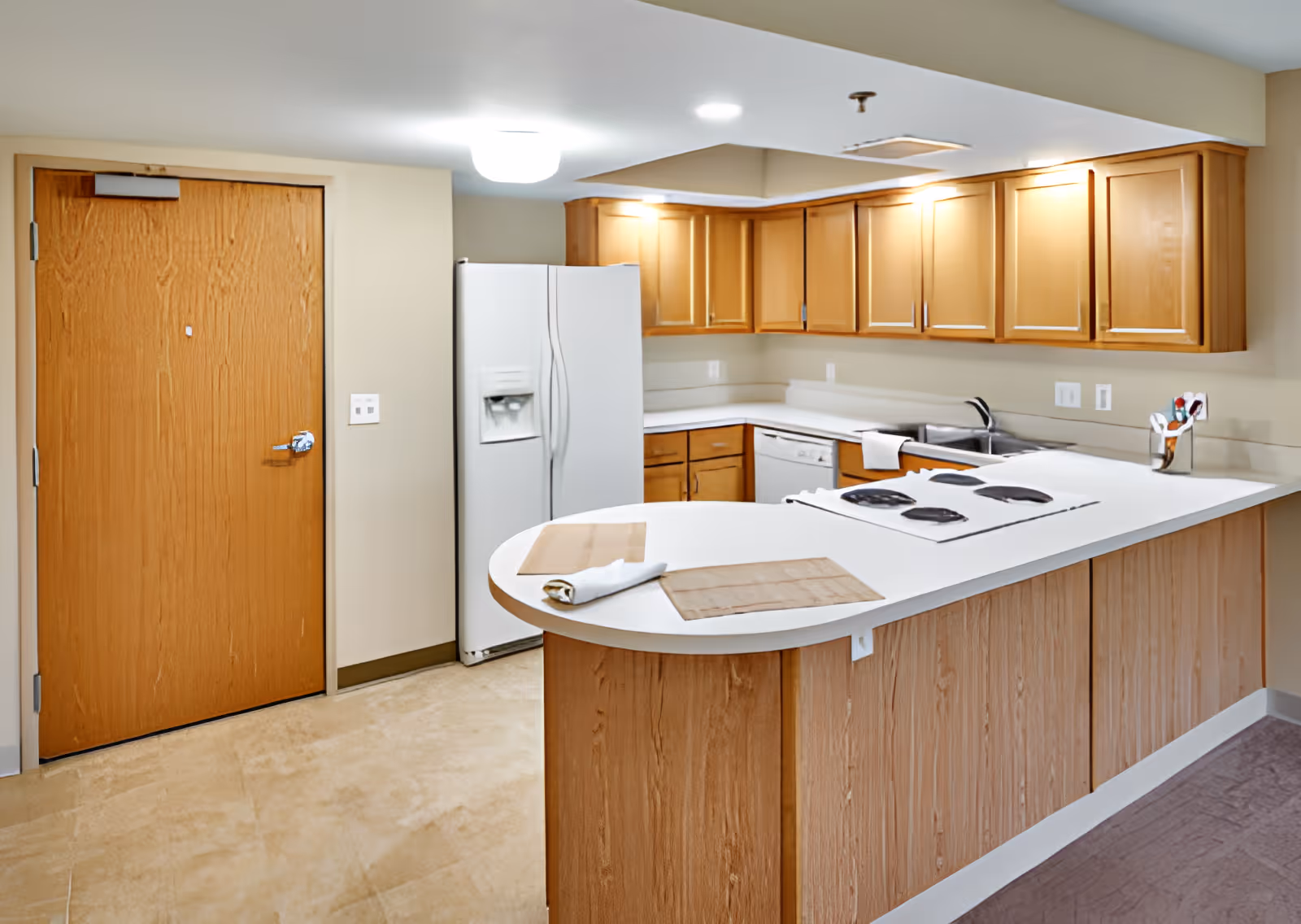 A clean and modern kitchen with wooden cabinets, a white refrigerator, a white electric stove on a curved countertop, a dishwasher, and a sink. The kitchen has beige walls and a wooden door on the left side.