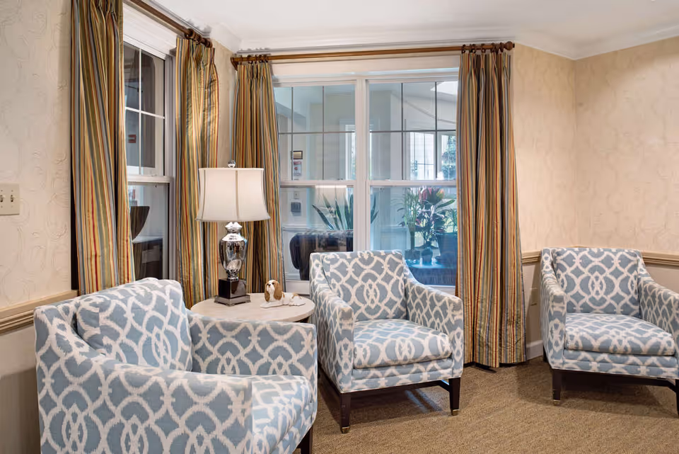 A cozy sitting area with three patterned armchairs arranged around a small round table with a decorative lamp and a small dog figurine. The room has beige wallpaper, striped curtains on the windows, and a carpeted floor.