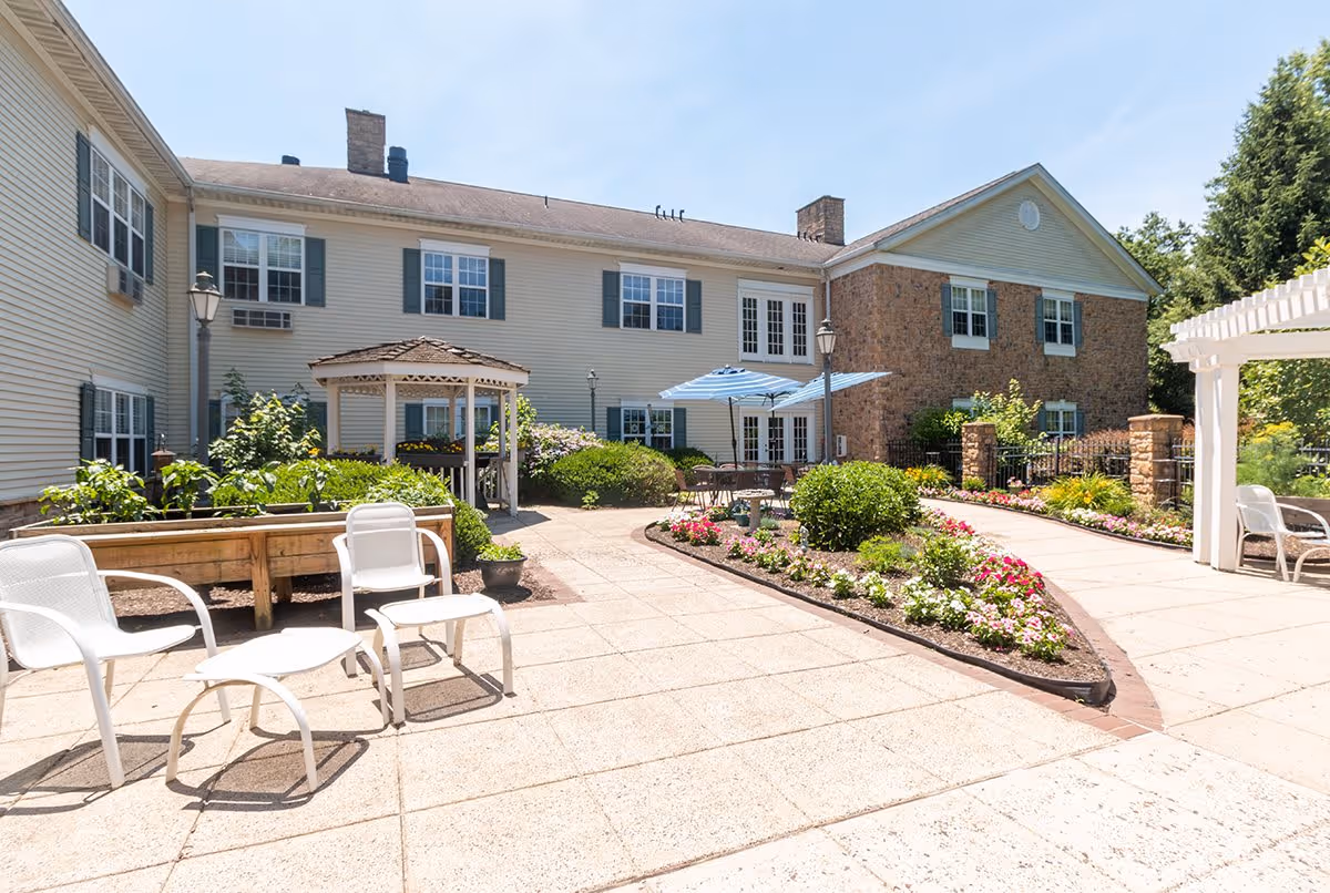 Outdoor courtyard area of a senior living facility with paved walkways, flower beds, white chairs, a small gazebo, and a table with a blue umbrella. The building in the background has beige siding and brick walls with multiple windows.