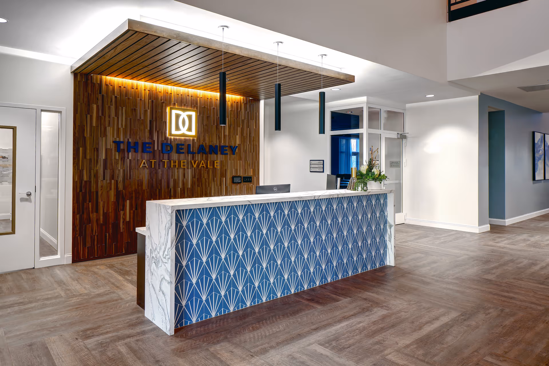 Reception area of The Delaney at the Vale featuring a modern front desk with a blue and white geometric pattern, a wood-paneled wall with the facility's illuminated logo and name, pendant lights hanging from a wooden ceiling accent, and a spacious floor with wood-style flooring.
