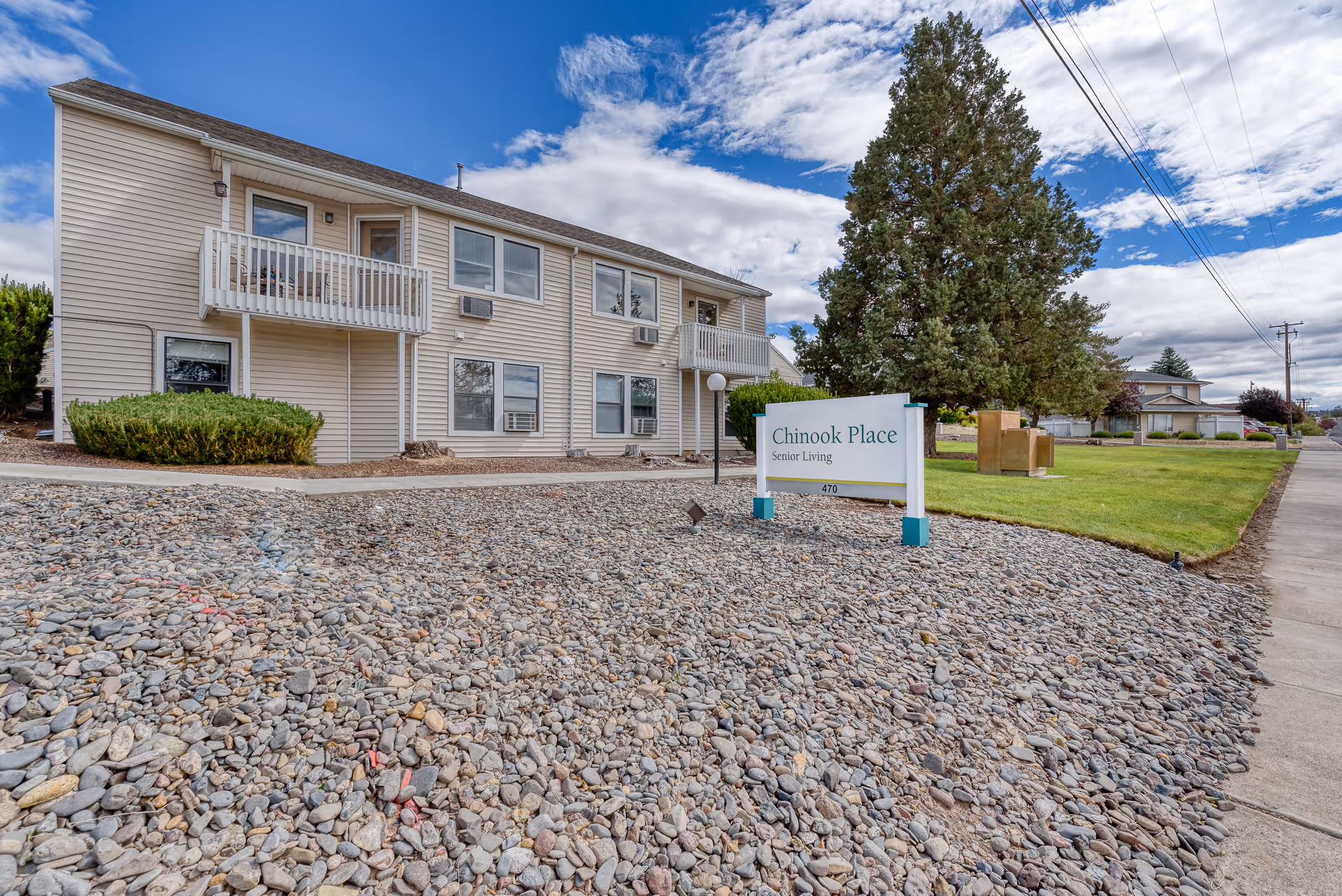 Two-story beige senior living building with a sign reading "Chinook Place Senior Living" in a rocky front yard and a large tree nearby.