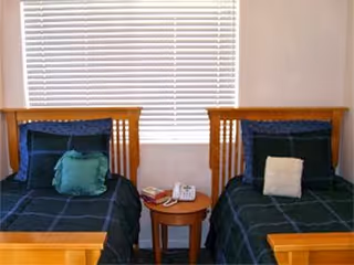 Two single beds with wooden headboards and green bedding separated by a small round nightstand beneath a window with closed blinds.