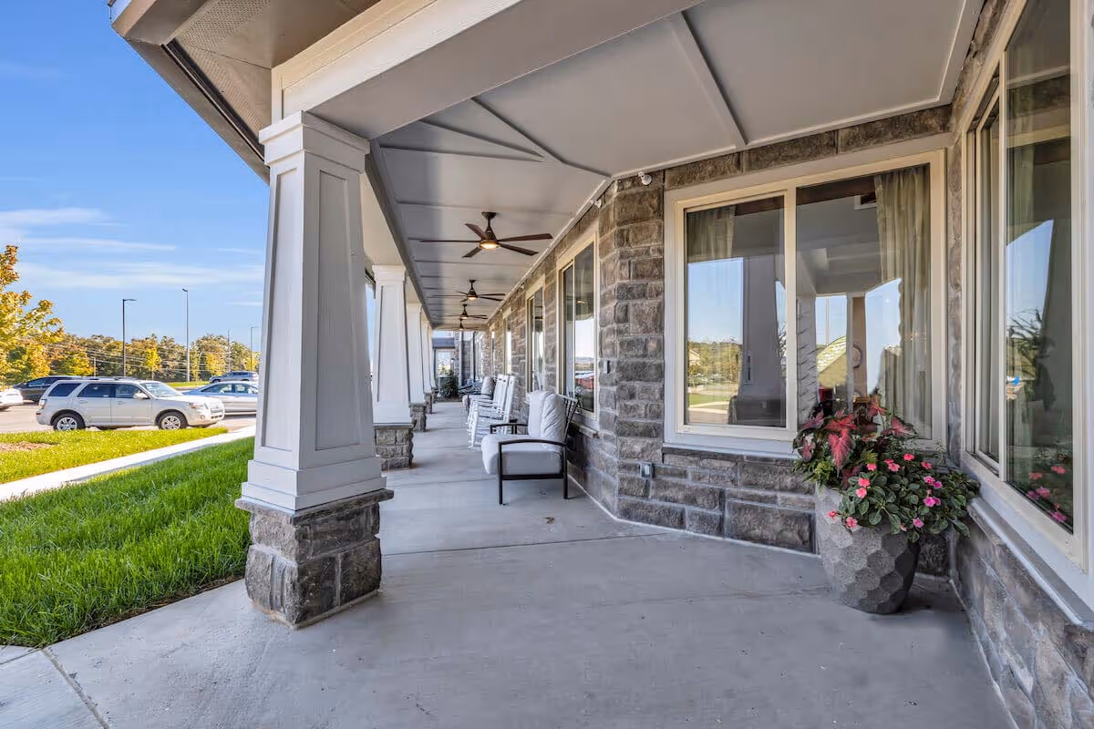 Covered outdoor walkway with stone pillars and ceiling fans, lined with cushioned chairs and a large planter with flowers, adjacent to a building with large windows and a parking lot visible in the background under a clear blue sky.