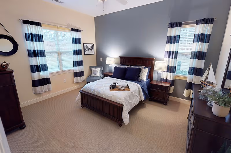 A cozy bedroom with a wooden bed frame, white and navy bedding, two nightstands with lamps, striped navy and white curtains on two windows, a cushioned chair with a pillow, a wooden dresser with decorative items including a model sailboat and a potted plant, and beige carpeted floor.
