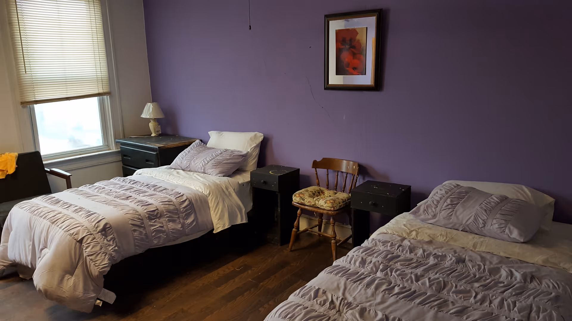 A bedroom with two single beds covered in light purple bedding. Between the beds are two small black nightstands with a wooden chair in the middle. A framed floral picture hangs on the purple wall above the chair. A window with closed blinds and a black chair with a yellow cloth are visible on the left side of the room.