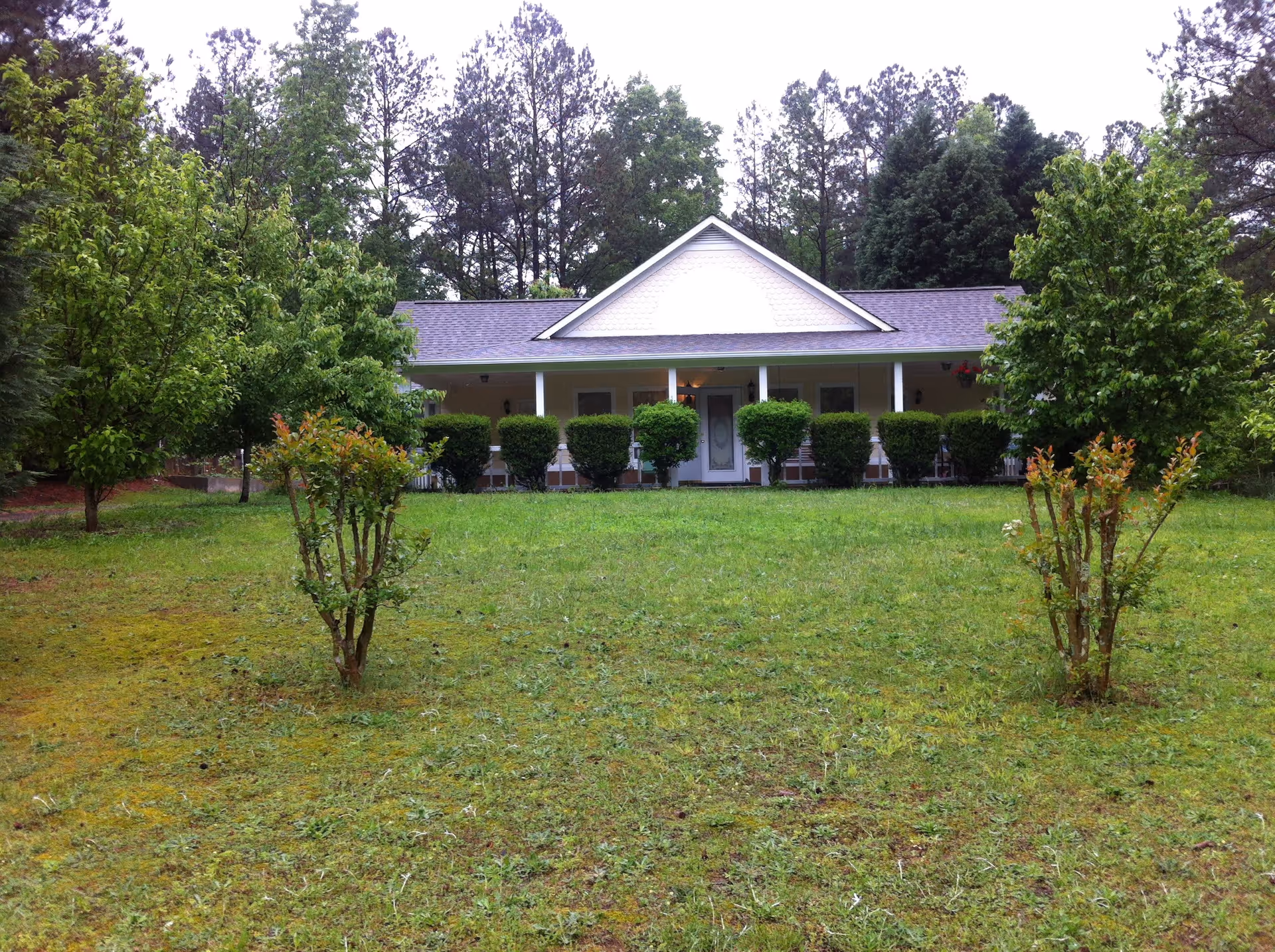 A single-story house with a front porch surrounded by neatly trimmed bushes and trees, set against a backdrop of tall pine trees and a grassy lawn in the foreground.