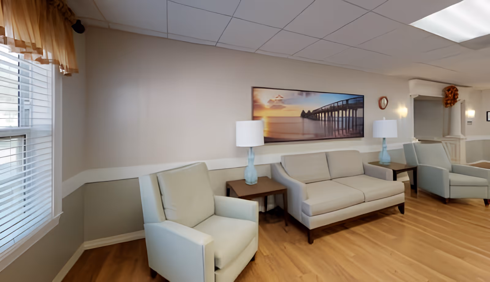 A bright seating area with sofas, armchairs, side tables and lamps beneath a wall landscape painting.