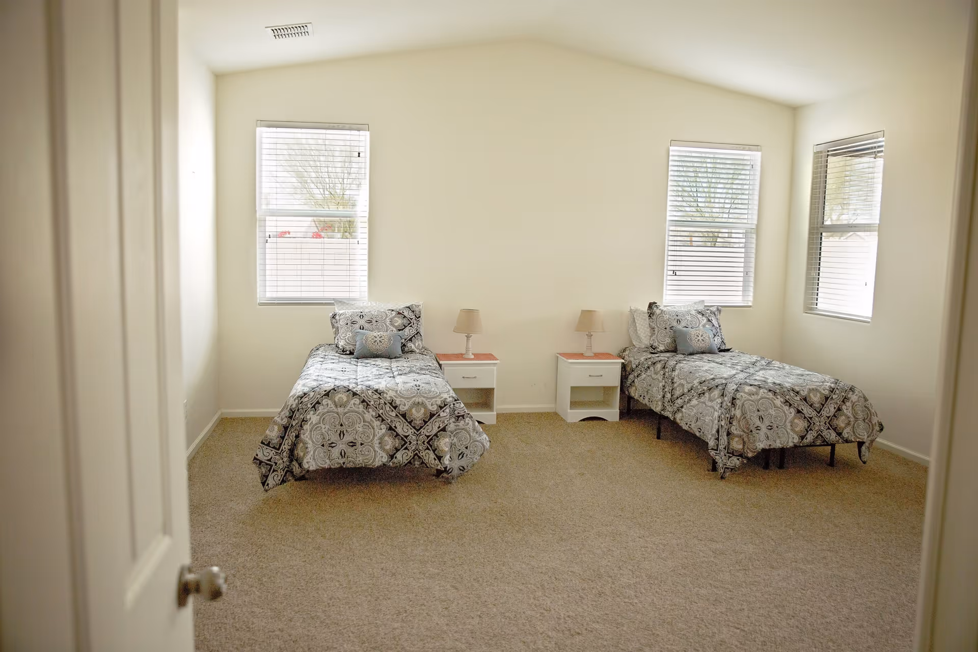 A simple bedroom with two twin beds separated by two white nightstands with lamps. Each bed has patterned bedding and pillows. The room has beige carpet and three windows with blinds letting in natural light.