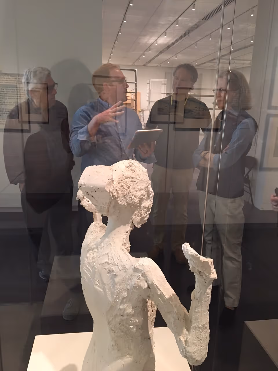 A white textured sculpture in a glass case with several people reflected and viewing it in a gallery interior.