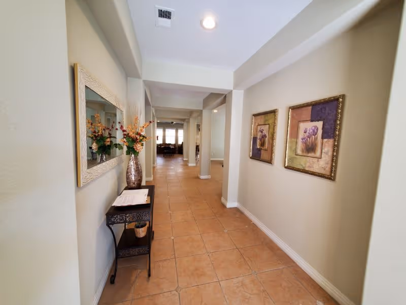 A long hallway with beige tiled floor and light-colored walls. On the left side, there is a small black table with a decorative vase holding flowers and a large mirror above it. On the right side, two framed floral paintings hang on the wall. The hallway leads to a room with windows letting in natural light.
