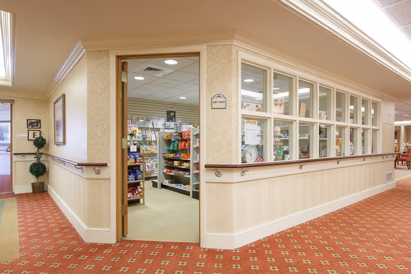 Interior view of a senior living facility hallway with beige walls and red patterned carpet. There is an open door leading to a small gift shop stocked with various items on shelves. The hallway has handrails along the walls and a potted plant near the corner.