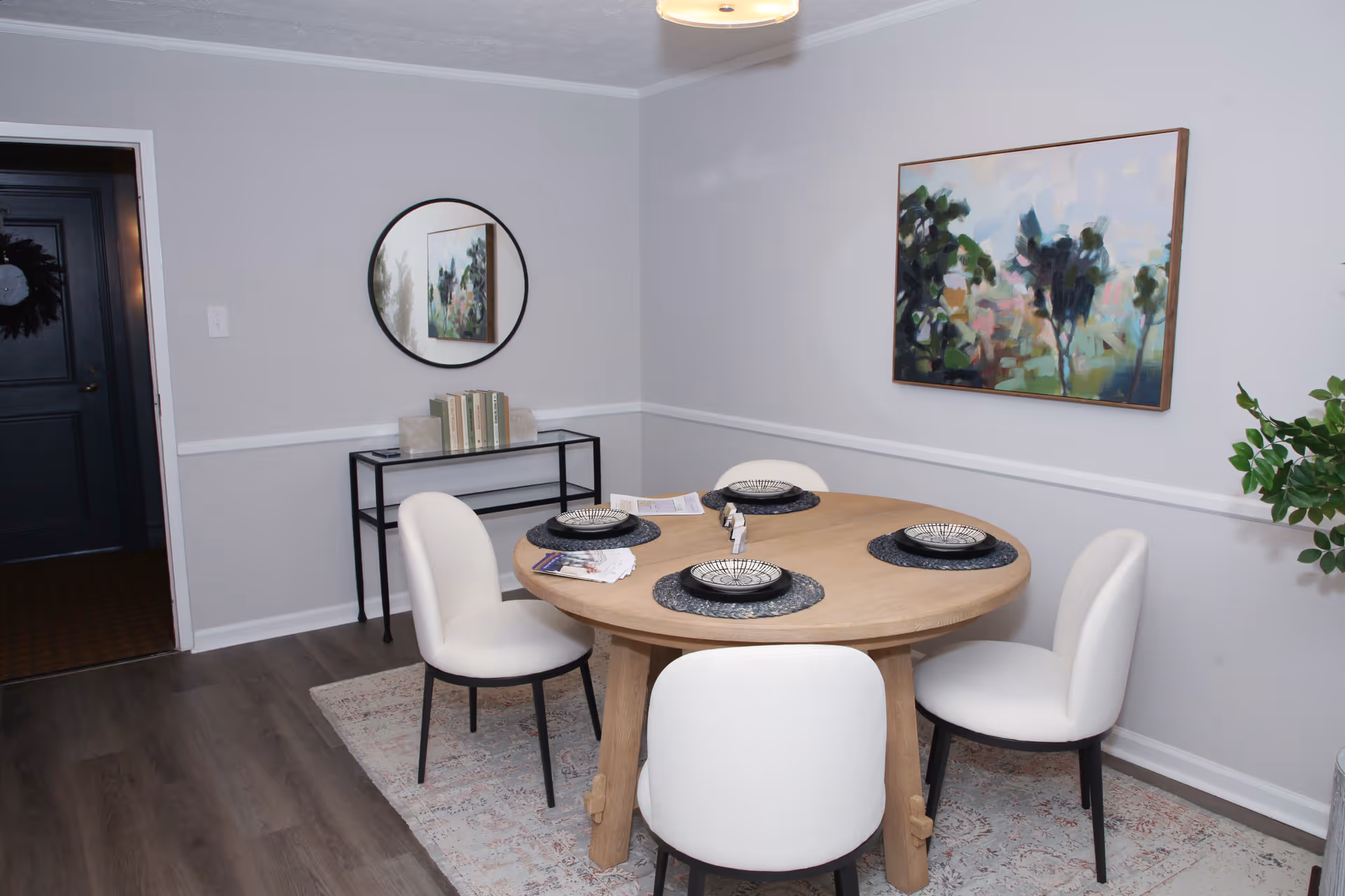 A dining room with a round wooden table set for four with black and white plates and black placemats. Four white cushioned chairs surround the table. A small black console table with books and decorative items is against the wall, beneath a round mirror. A colorful abstract painting hangs on the wall, and a green plant is visible on the right side. The floor is wood with a patterned rug under the table.