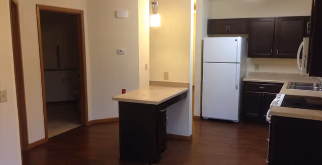 Interior view of a kitchen area with dark wood cabinets, a white refrigerator, a stove, a microwave, and a small island with a countertop. The floor is wooden, and there is a doorway leading to another room.