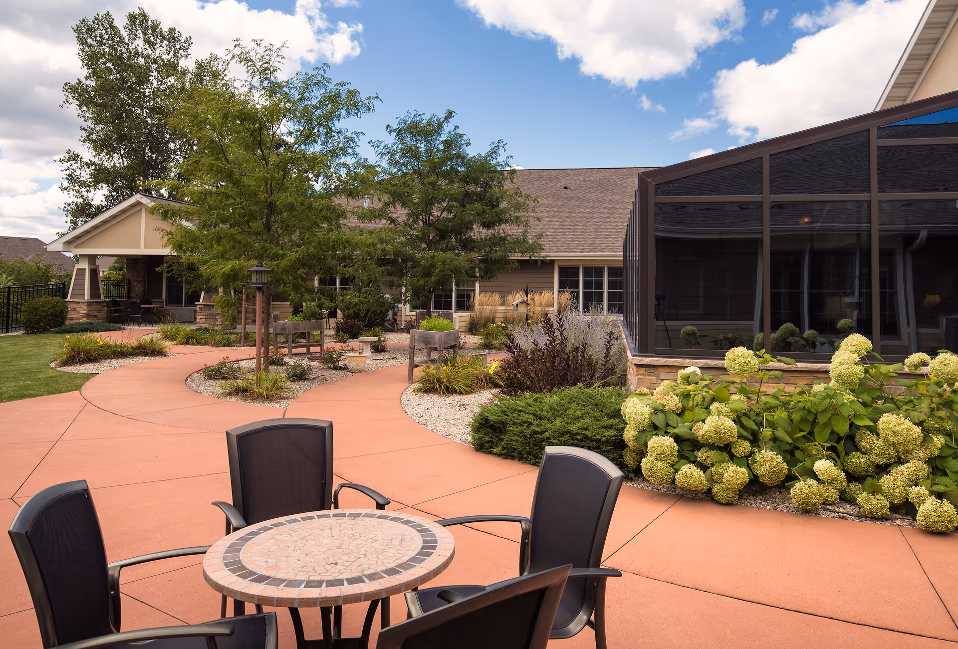 Outdoor patio area at Mission Creek Senior Living with a round mosaic table and four black chairs in the foreground. The patio is surrounded by landscaped garden beds with green shrubs, flowering plants, and trees. A building with large windows and a screened-in porch is visible in the background under a partly cloudy sky.