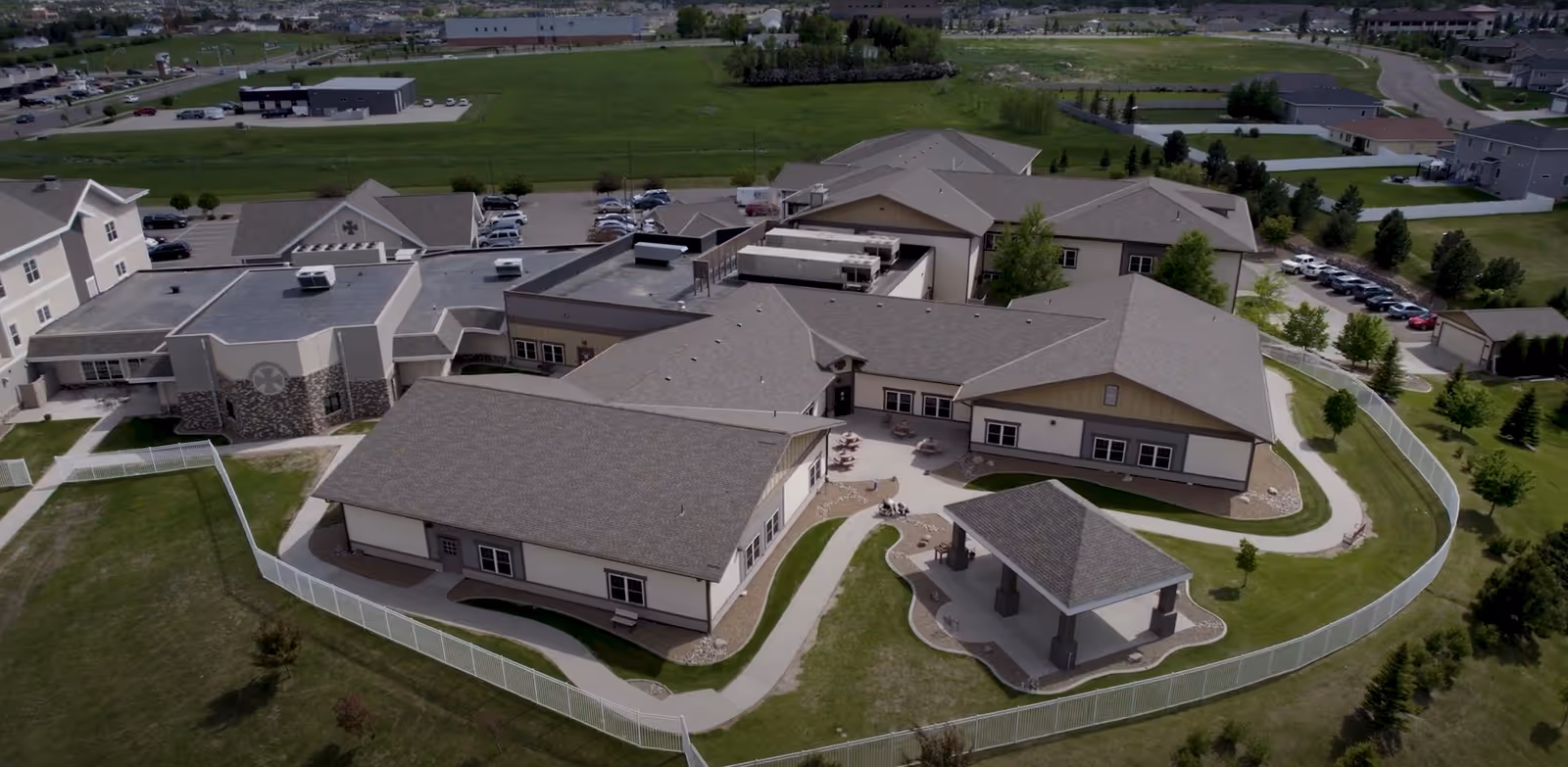Aerial view of Benedictine Living Community-Bismarck, showing a large, single-story building complex with multiple connected sections, surrounded by a white fence and green lawns. There is a small covered pavilion area and several outdoor seating areas. Parking lots and additional buildings are visible in the background.