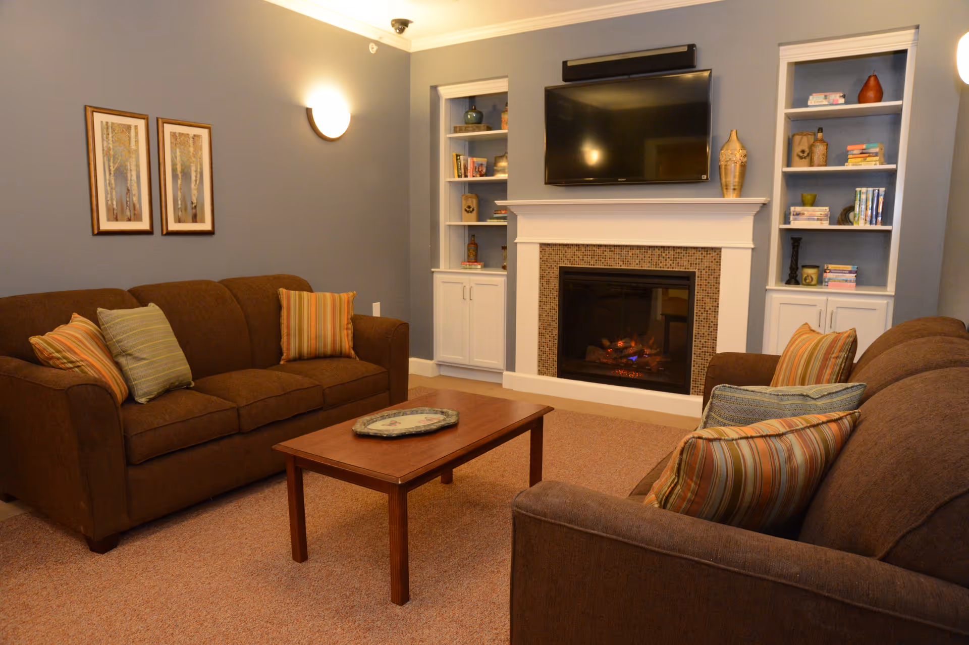 A cozy living room with two brown sofas adorned with striped and solid pillows facing a wooden coffee table. The room features a fireplace with a mosaic tile surround and a mounted flat-screen TV above it. Built-in white shelves flank the fireplace, holding books and decorative items. The walls are painted blue, and two framed pictures hang on one wall.