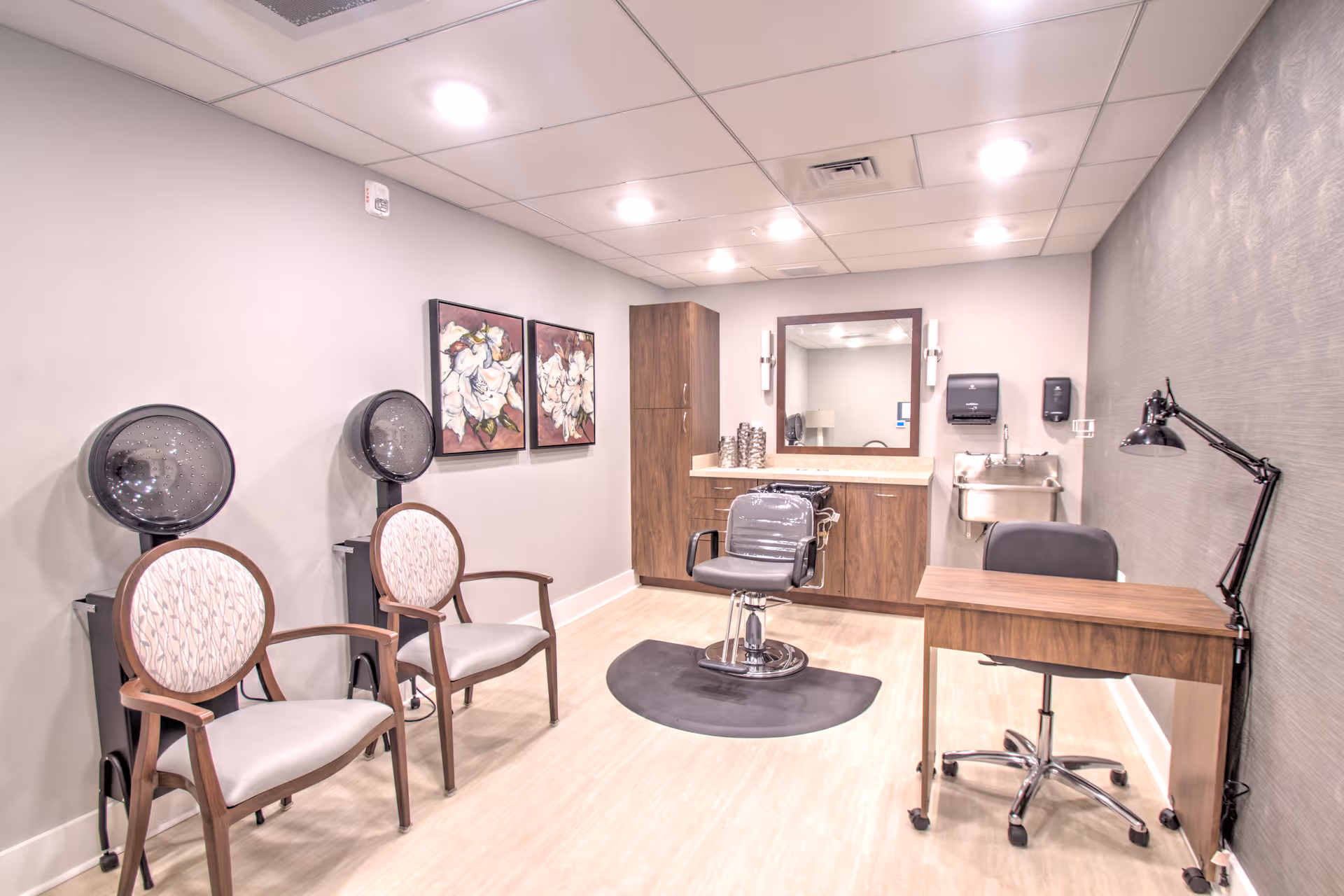 A well-lit salon room with two vintage hair dryers and chairs on the left, a styling chair in front of a large mirror and wooden cabinetry, a small sink on the right wall, and a wooden desk with a black adjustable lamp and office chair.