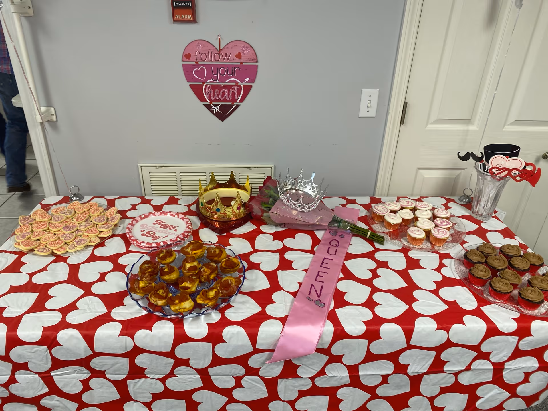 A table covered with a red tablecloth with white hearts is set up with various desserts including heart-shaped cookies, cupcakes with pink and brown frosting, and a plate of pastries. On the table are two crowns, a pink sash labeled 'QUEEN', and a bouquet of flowers. A heart-shaped wall decoration above the table reads 'follow your heart'.