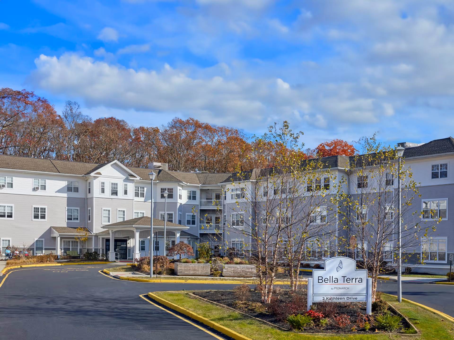 Exterior view of Monarch Bella Terra senior living facility showing a large, multi-story building with white and gray siding, surrounded by trees with autumn foliage under a partly cloudy blue sky. There is a driveway leading to the entrance and a sign in front that reads 'Bella Terra by Monarch, 2 Kathleen Drive.'