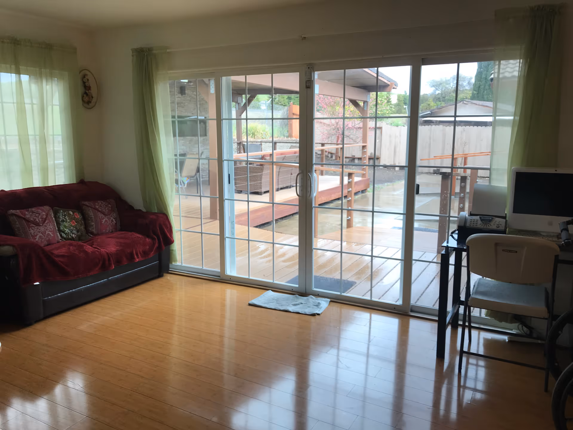 Interior view of a living room with a red couch covered with a red throw and decorative pillows on the left, a desk with a computer and chair on the right, and large sliding glass doors in the center leading to an outdoor patio area with wooden decking and outdoor furniture.