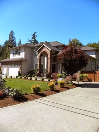 Large two-story suburban house with a landscaped front yard, driveway, and garage under a clear blue sky.