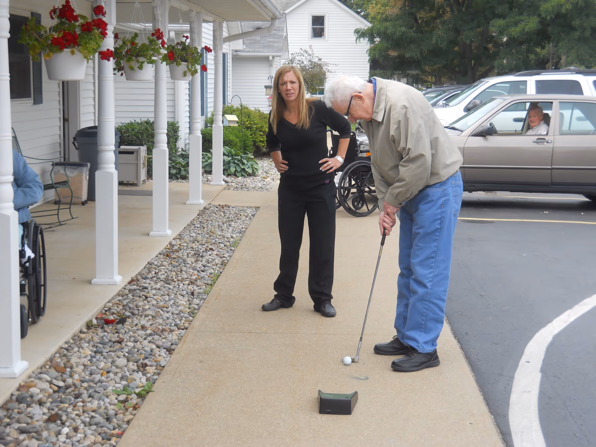 An elderly man is playing mini-golf on a sidewalk outside a building, with a woman standing nearby watching him. There are hanging flower pots with red flowers on the porch to the left, and parked cars in the background. Another person is visible sitting in a wheelchair on the left side of the image.