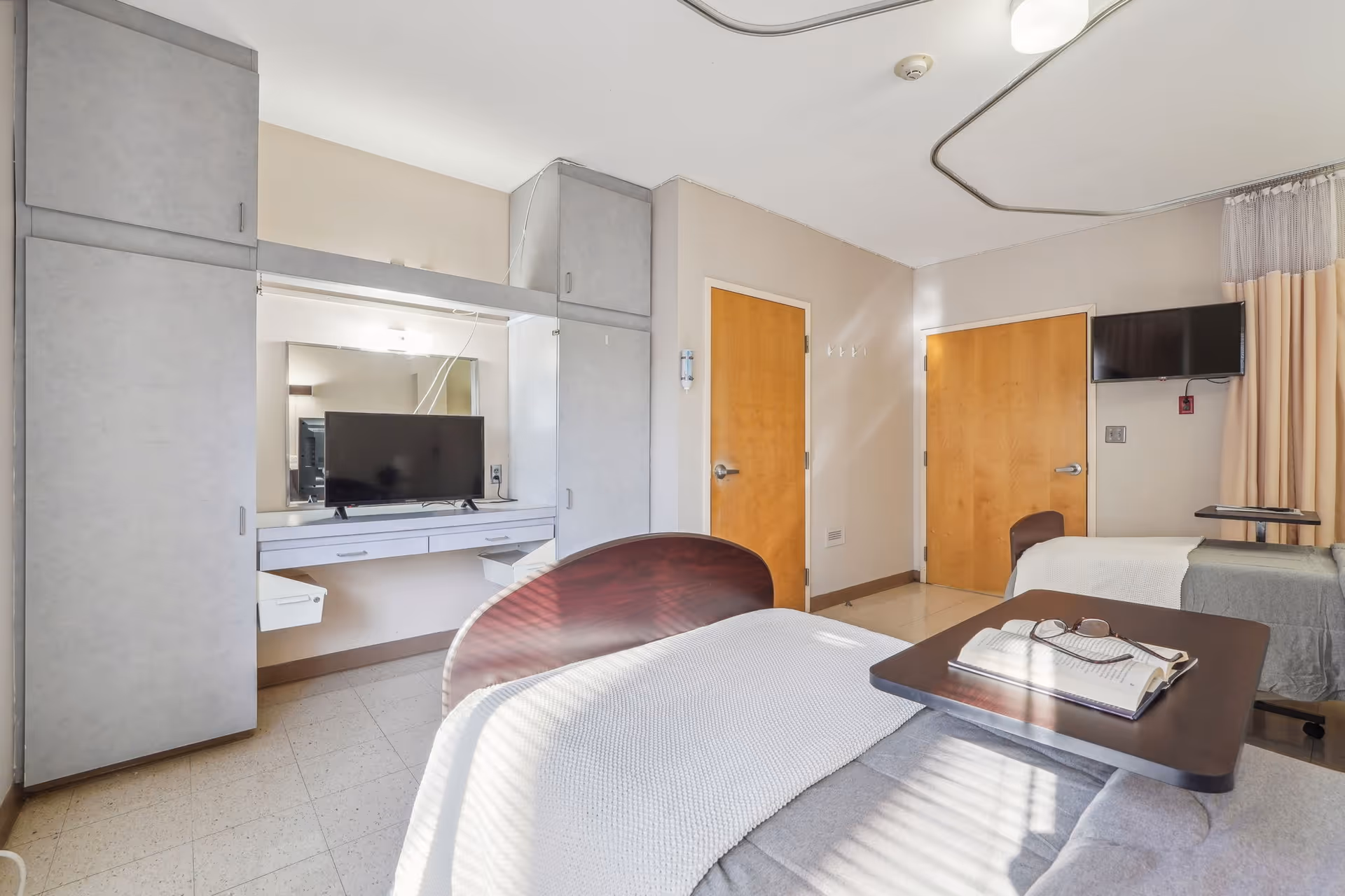 A bright and clean room with two single beds, each with a white blanket. One bed has a small table with an open book and glasses on it. There is a flat-screen TV mounted on the wall near one bed and another TV on a desk with built-in cabinets and drawers. The room has two wooden doors, beige walls, and a curtain partially drawn to the side.