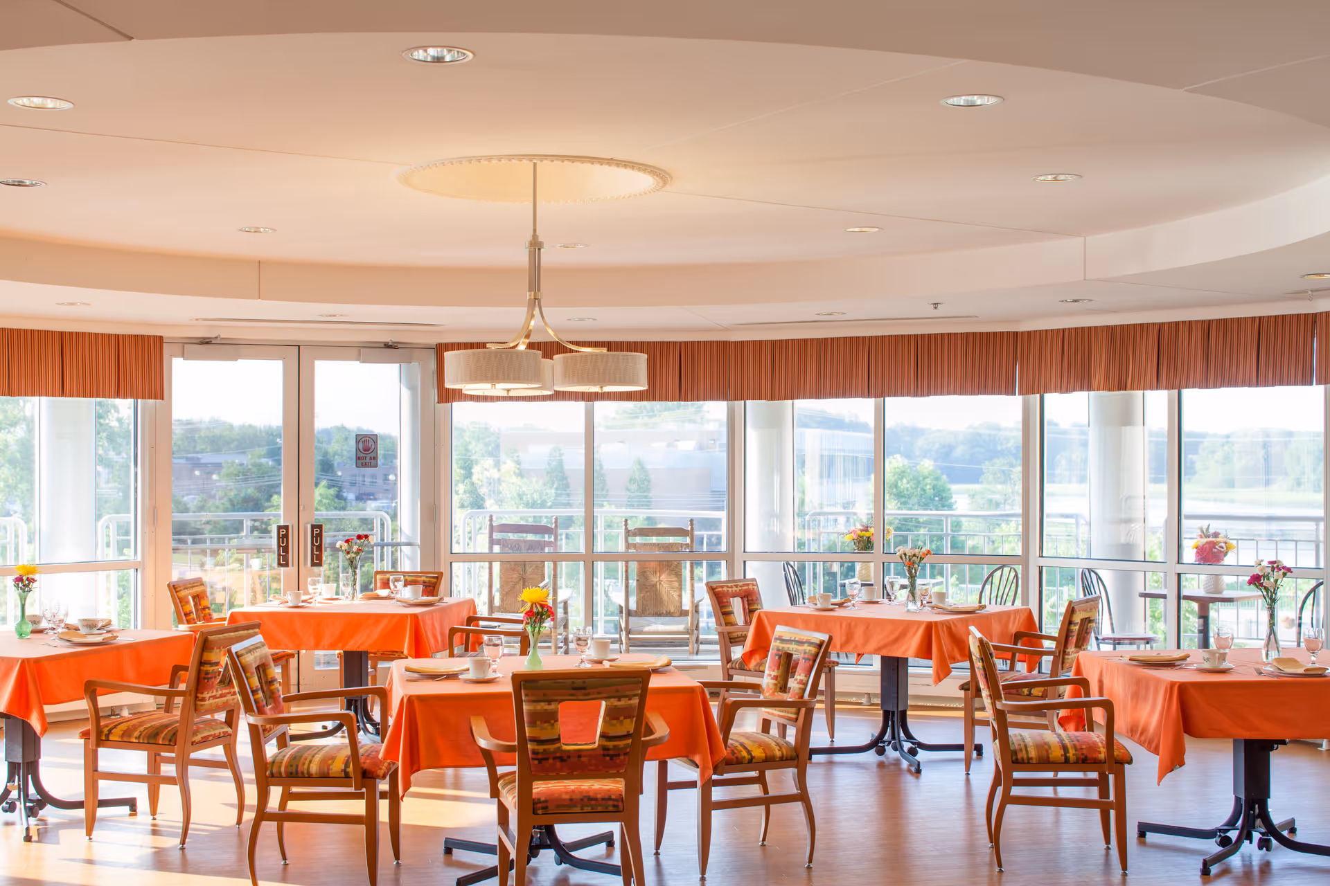 A bright dining room with several tables covered in orange tablecloths, each set with cups, plates, and small flower vases. Wooden chairs with patterned cushions surround the tables. Large windows and glass doors let in natural light and offer a view of greenery outside. A modern ceiling light fixture hangs above the center of the room.