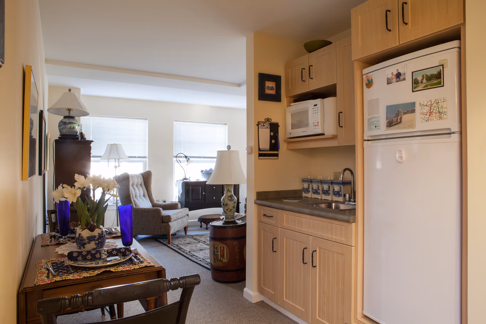 A cozy interior space featuring a small kitchen area with light wood cabinets, a white refrigerator adorned with magnets, a microwave, and a countertop with a sink and decorative canisters. Adjacent to the kitchen is a living area with a cushioned armchair, a side table with a lamp, and a window with blinds letting in natural light. A wooden table with blue glass vases and a floral arrangement is also visible in the foreground.