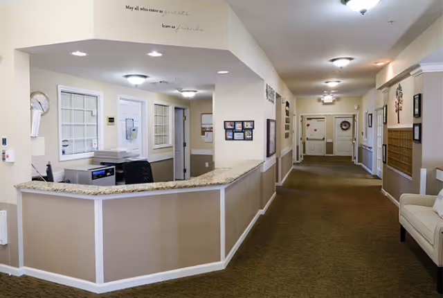 Reception area with a curved granite countertop desk, office chair, and computer equipment. The hallway extends to the right with beige walls, carpeted floor, framed pictures, and a cushioned bench. Ceiling lights illuminate the space. A wall decal above the reception desk reads 'May all who enter as guests leave as friends.'