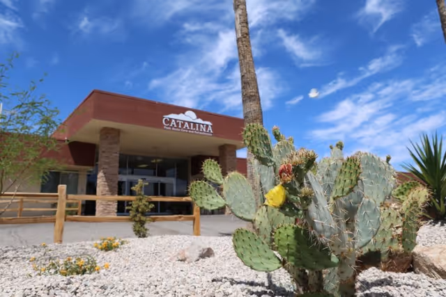 Exterior view of Catalina Post Acute And Rehabilitation facility with a clear blue sky, desert landscaping featuring a cactus with a yellow flower, and a building entrance with stone pillars and a wooden fence.