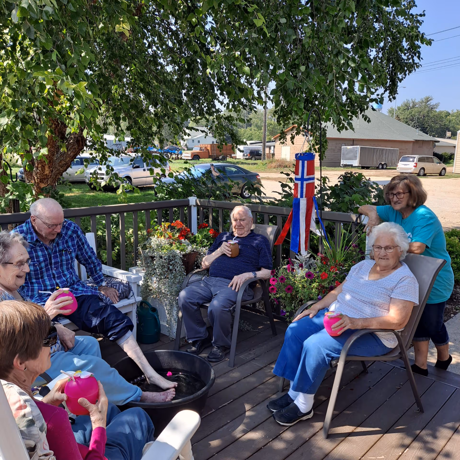 A group of elderly residents and a caregiver socializing on a shaded outdoor deck with potted flowers and a foot basin.
