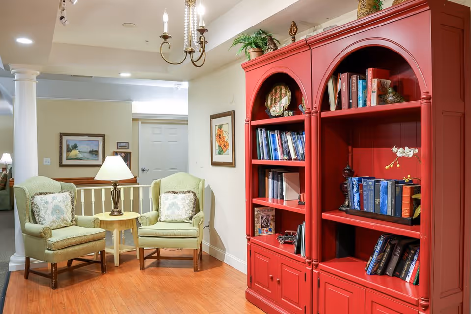 A cozy sitting area in a senior living facility featuring two green upholstered armchairs with patterned cushions, a small wooden side table with a lamp between them, and a large red bookshelf filled with books and decorative items. The room has wooden flooring, framed artwork on the walls, and a chandelier hanging from the ceiling.