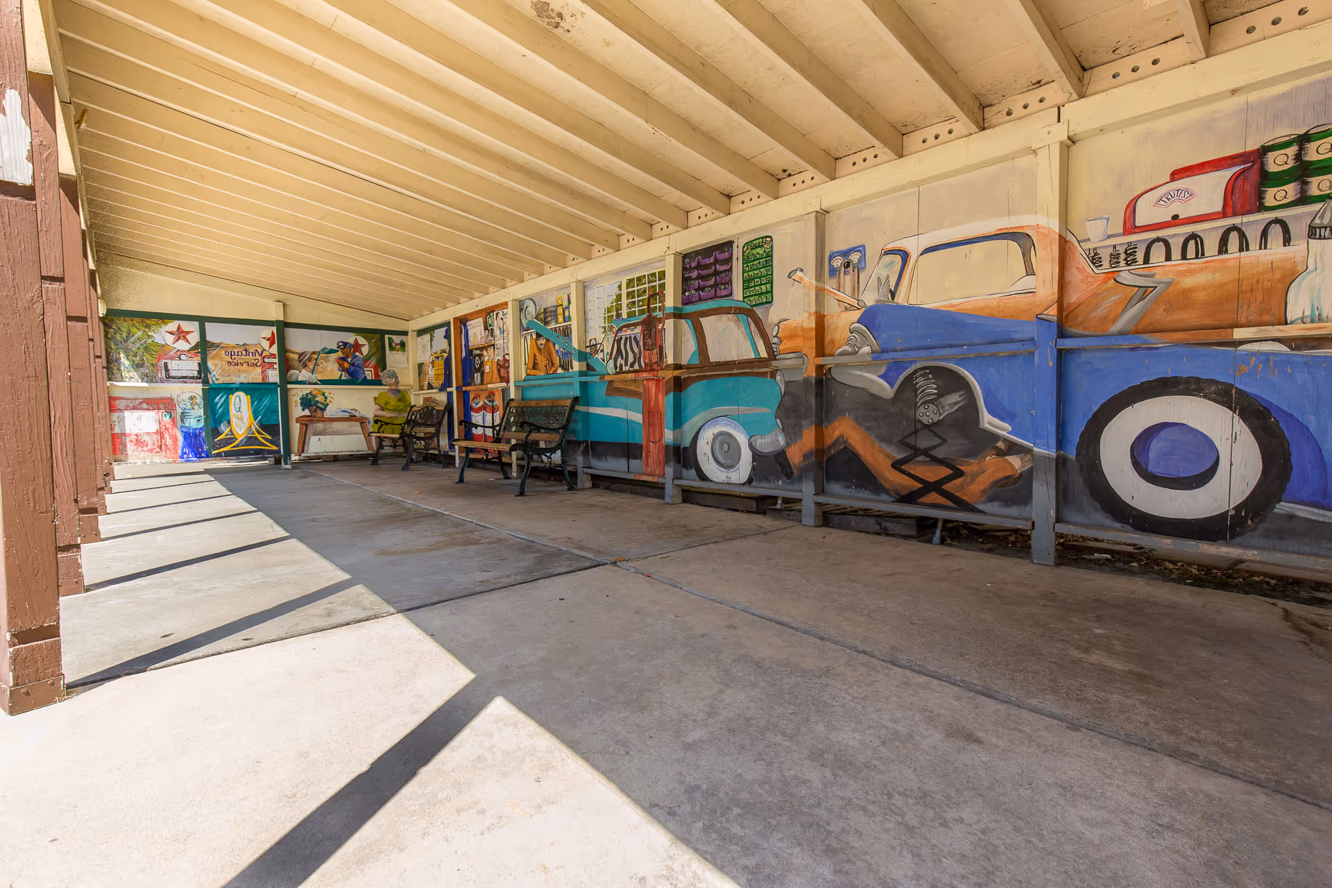 Covered outdoor walkway with a sloped ceiling, benches, and a colorful mural of vintage cars painted on the wall.