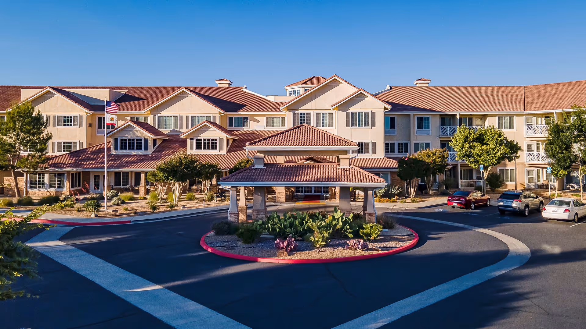 Front exterior view of Holiday Rancho Village, a large multi-story senior living facility with beige walls and red-tiled roofs. There is a circular driveway with a covered entrance in the center, landscaped with desert plants and trees. Several cars are parked along the driveway, and two flagpoles with American and California state flags are visible near the entrance.