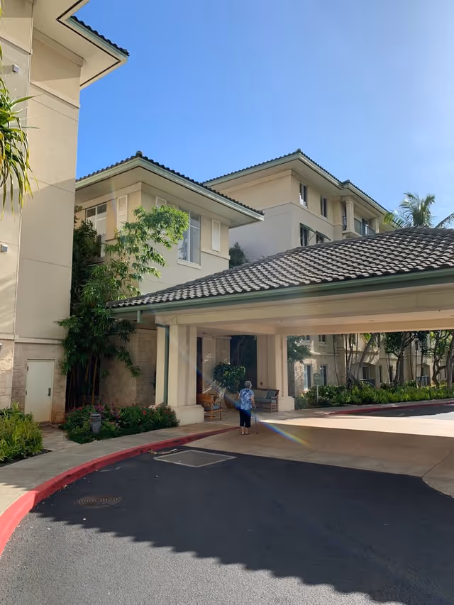 Covered entrance and drive at a multi-story residential building with landscaping and a person using a cane near the drop-off.
