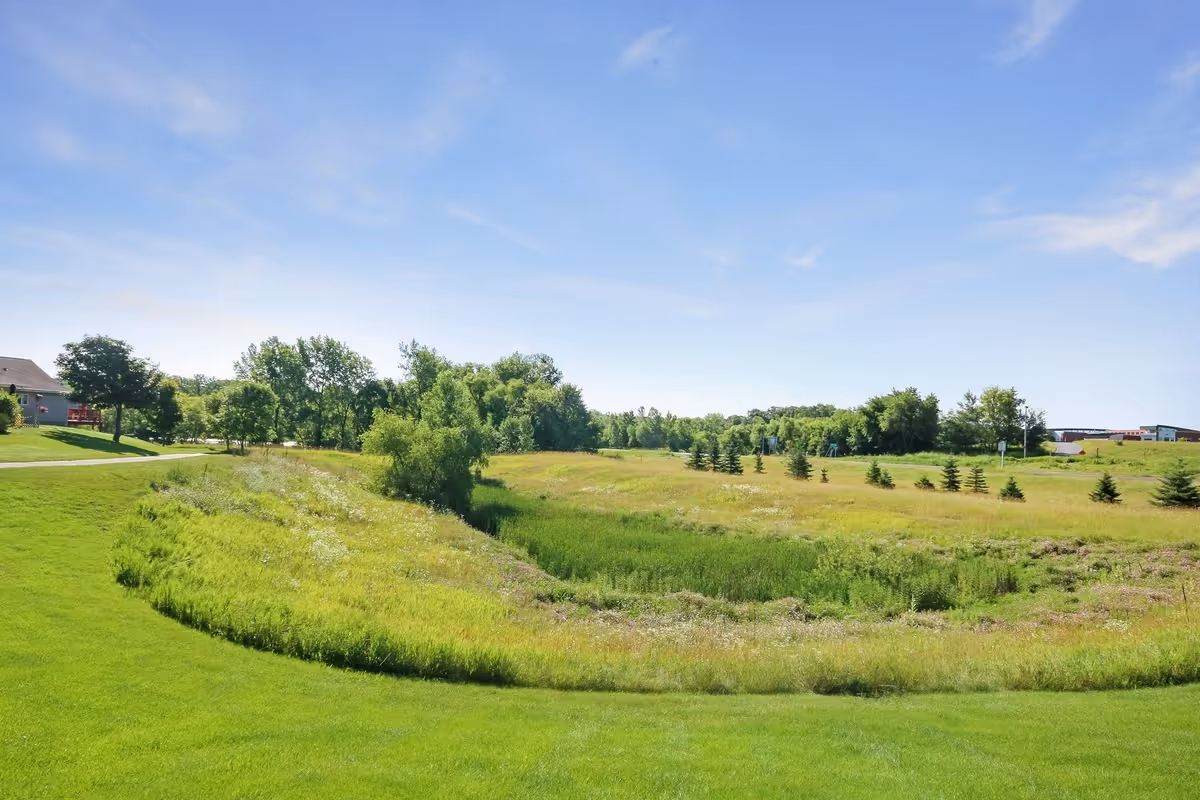 A wide, open grassy field with a variety of green trees and shrubs under a clear blue sky with a few wispy clouds. There is a paved path on the left side and some buildings visible in the distance on the right.