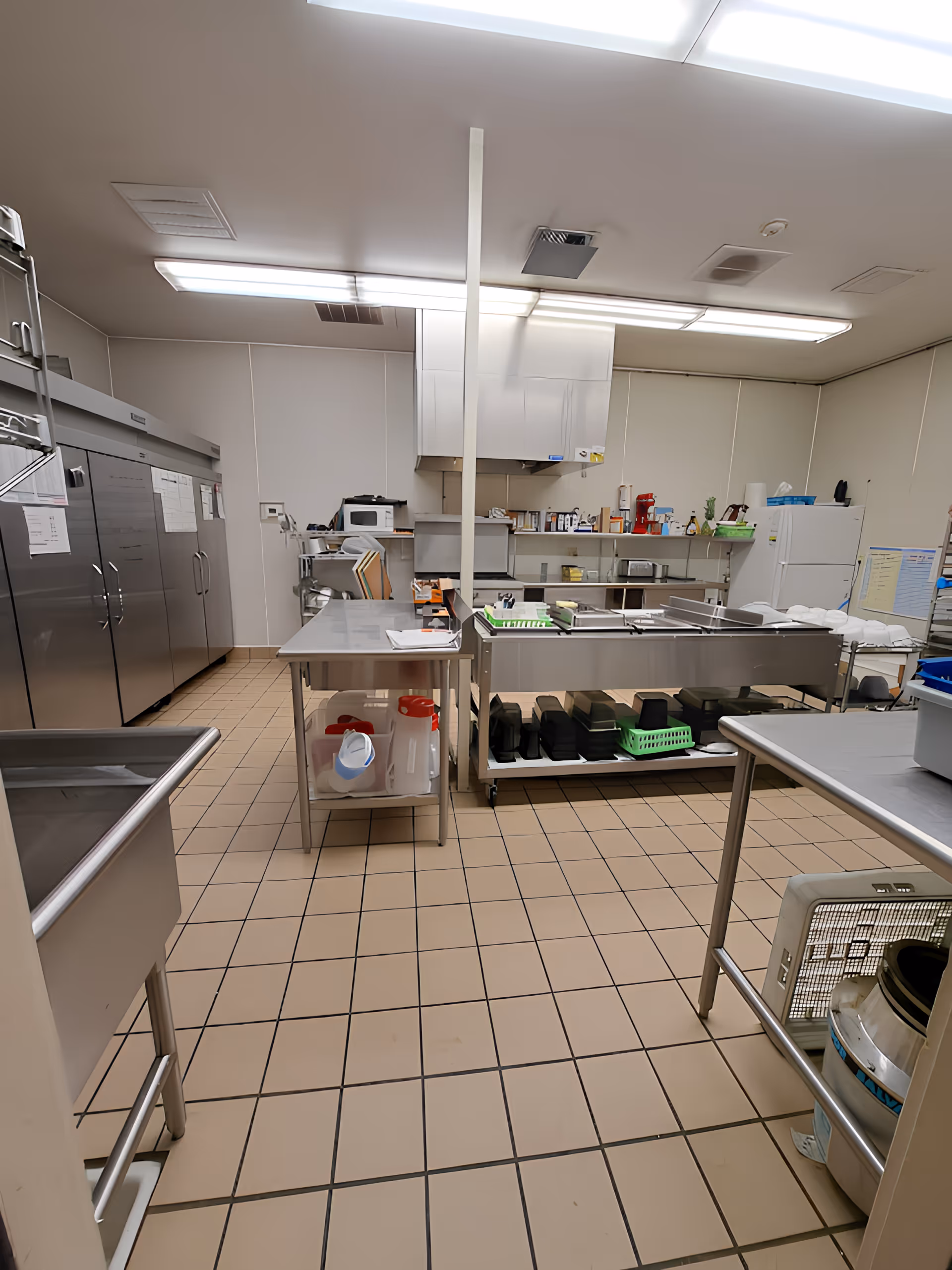A commercial kitchen with stainless steel appliances and work tables. There are large refrigerators on the left side, a microwave on a counter in the back, and various kitchen supplies and containers on shelves and tables. The floor is tiled with beige square tiles, and the ceiling has fluorescent lighting.
