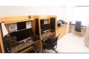 A small computer room with three workstations, each equipped with a desktop computer, monitor, and office chair. The room has light-colored walls and flooring, with a window covered by a sheer curtain allowing natural light to enter.