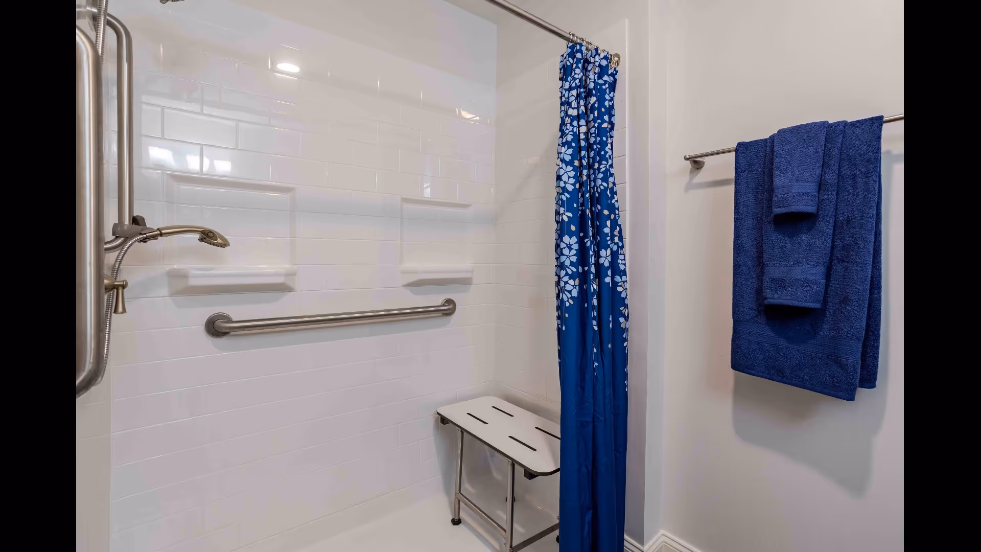 A bathroom shower area with white tiled walls, a metal grab bar, a handheld showerhead, a foldable shower seat, and a blue shower curtain with white floral patterns. On the right wall, there is a towel rack holding two dark blue towels.
